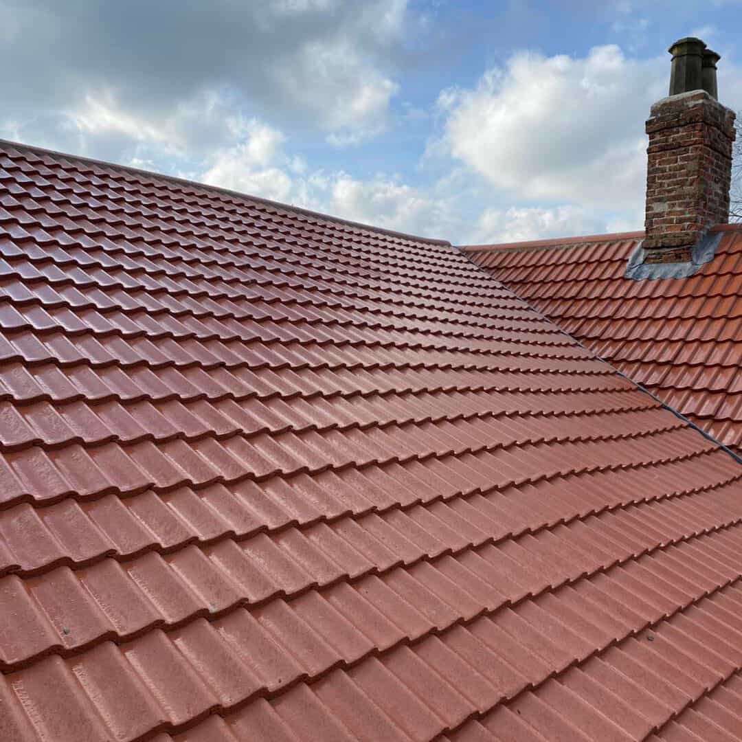 A red tiled roof with clean, evenly arranged tiles and a brick chimney, set against a partly cloudy blue sky.